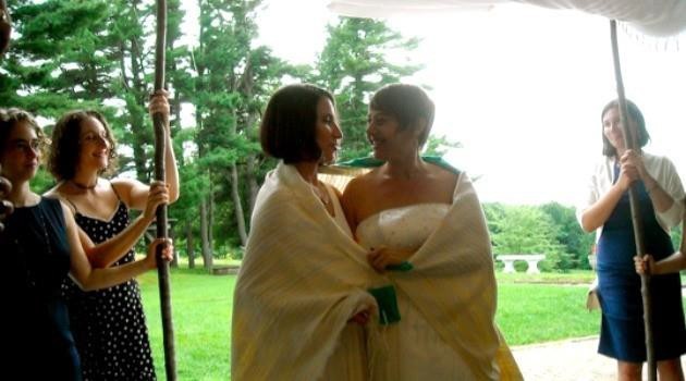 Image is of two smiling women - Idit Klein and Jordan Namerow - standing under a chuppah at their wedding.