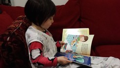 Image is of a child reading a picture book. The book is a copy of the Purim Superhero.