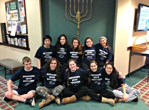 Image is a group of 9th grade students sitting together in front of a Menorah. They're wearing black t-shirts that read "Buddies Note Bullies"