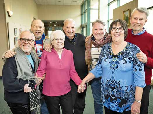 A group of LGBTQ elders standing together, smiling and holding hands. 