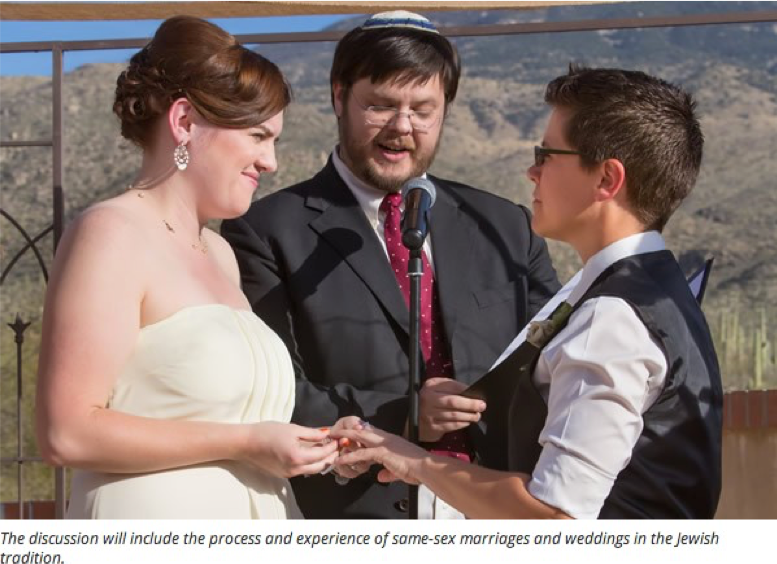 Image of two women getting married standing in front of a rabbi. One woman is wearing a white dress and the other is wearing a suit. They are holding hands.