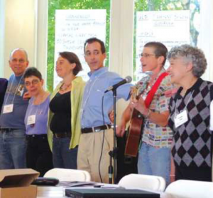 Image of people standing with their arms around each other, singing. A man is holding a guitar and is singing into a microphone.