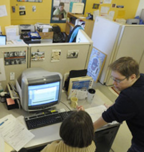 Image of two people. One is sitting at a desk in an office space looking at a computer. The other person is standing next to the desk talking to the firs person.