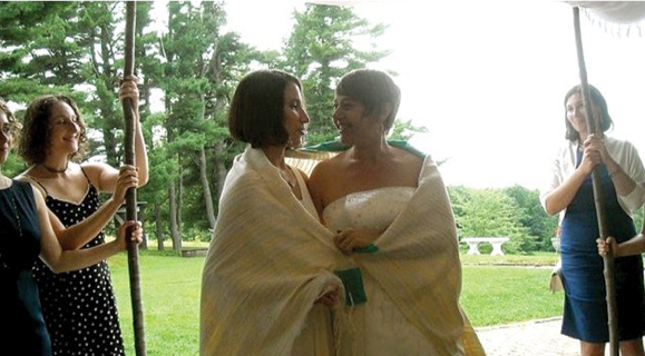 Two smiling women in wedding dresses standing under a huppah.