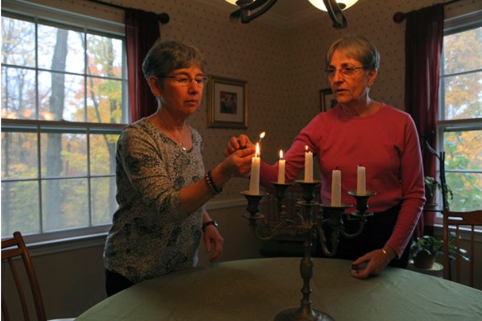 Image of Anne Corey and Connie Knapp lighting a menorah in their dining room.