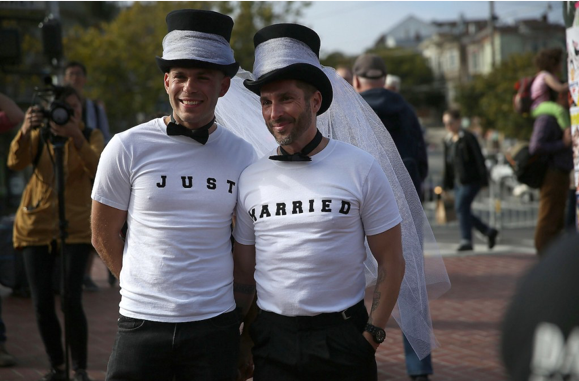 Image of two men standing outside wearing white shirts and black top hats with white veils on them. One man's shirt says "Just"; the other man's shirt says "Married."