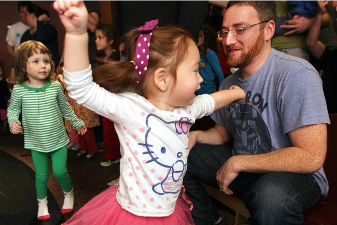 Image of a young girl in a pink tutu and Hello Kitty shirt dancing in front of her father.