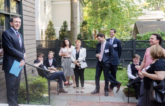 Image of a group of people sitting and standing outside. Matt Nosanchuk is on the far left, wearing a suit and smiling.