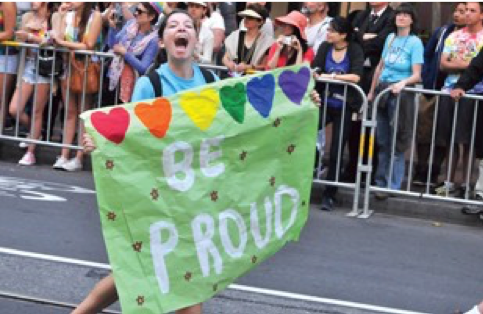 Image of a person on a street holding a giant green sign with rainbow hearts that says "Be Proud."