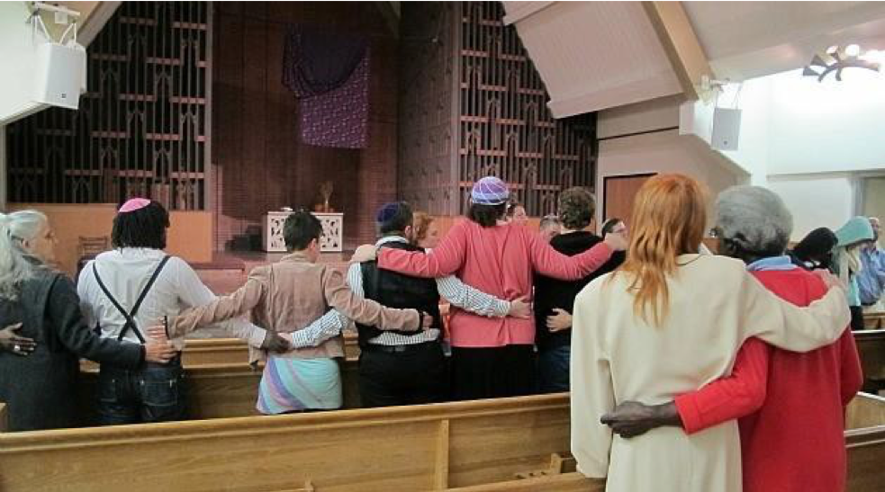 Image of a group of people standing in a synagogue with their arms around each other.