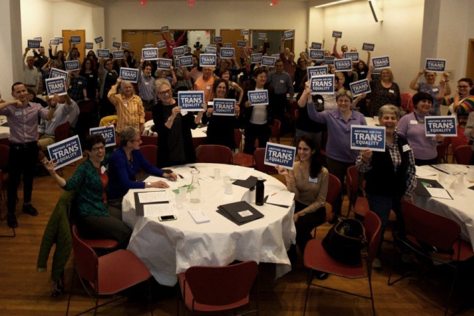 Image of a room full of people standing and sitting around tables. They are all holding blue signs that read: "Another Jew for Trans Equality"