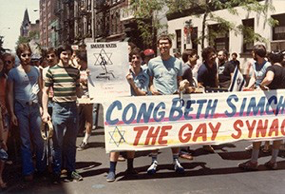 Image of a crowd of people at a Pride Parade, holding a sign with the words "CongBeth Simch" visible -- under that, "The Gay Synagogue" is written.