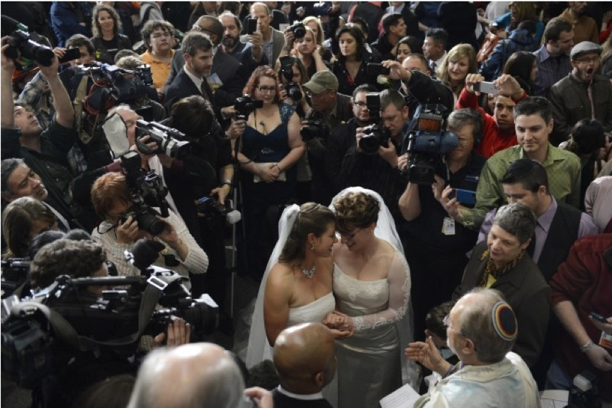 Image of two women in wedding dresses surrounded by photographers and cameras.