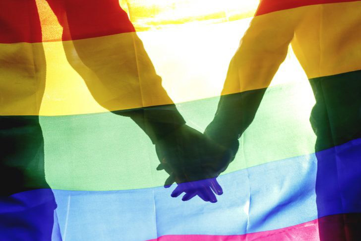 Picture of two intertwined hands on a backdrop of a rainbow LGBTQ flag.