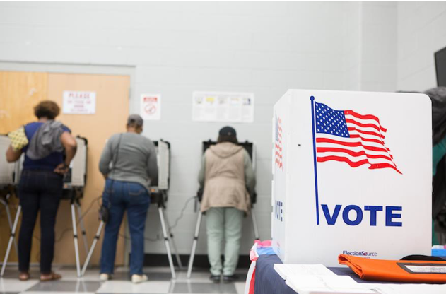 Image of three people voting in a polling station. A sign with the American flag and the word "VOTE" is visible.