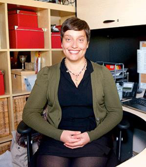 Image of Rebecca Cariati sitting at her desk with her hands folded in her lap, smiling at the camera.