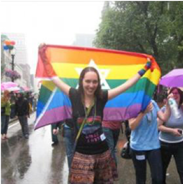 Image of a woman standing in the rain with a rainbow Pride flag with the Star of David on it. The person is proudly holding up the flag.