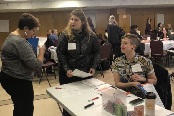 Image of two people behind a desk talking to another person standing in front of the desk in a room full of tables. 