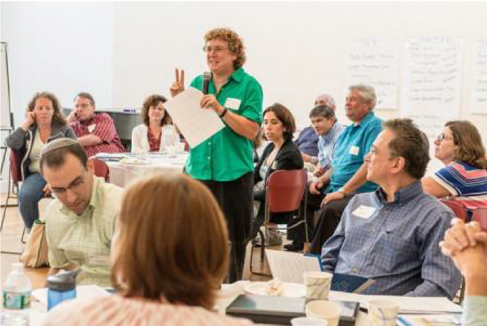 Image of a person in a green shirt standing in the middle of people sitting around tables. The person is holding a microphone and speaking to the crowd.