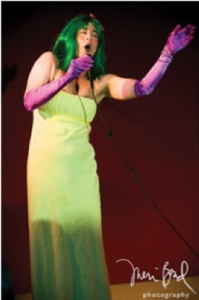 Image of a person in a green dress, green wig, and long purple gloves singing into a microphone.