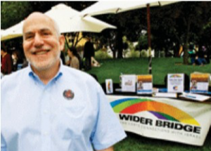 Image of Arthur Slepian standing outside in front of a table with a banner that reads: "A Wider Bridge"