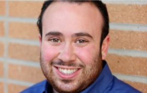 Image of Patrick Rock in front of a brick wall. He is wearing a blue shirt and is smiling.