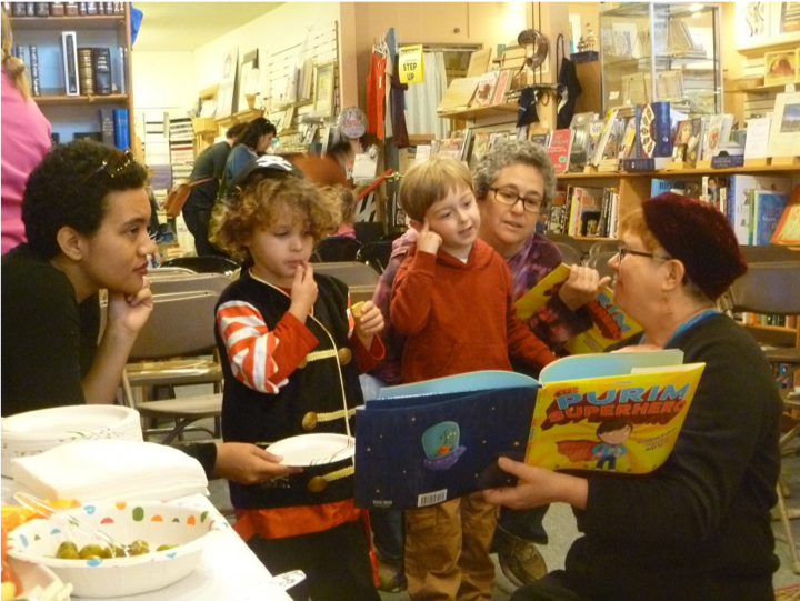 Image of Elisabeth Kushner reading "The Purim Superhero" to two kids. Two adults look on.