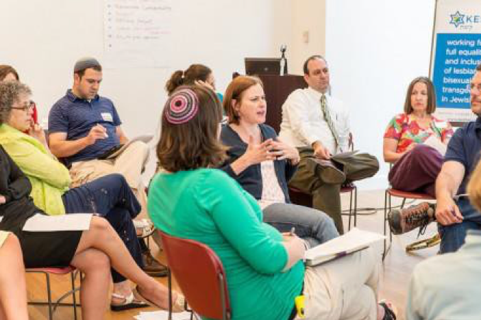 Image of a group of people sitting in a circle in chairs, listening to a person in the middle speak.