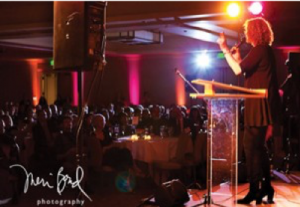 Image of a person speaking at a podium in front of a crowd of people. The lights are dimmed.