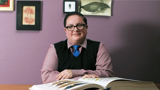 Image of Rabbi Elliot Kukla sitting with a large book in front of a purple wall. He is wearing a pink shirt, a blue tie, and a black vest.