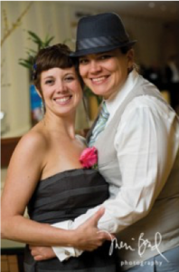 Image of two people with their arms around each other, smiling. The person on the left is wearing a black dress. The person on the right is wearing a white shirt with a tan vest and a fedora.