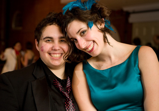 Image of two people sitting together smiling. The person on the left is wearing a suit and the person on the right is wearing a teal dress with matching feathers in her hair.