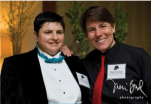 Image of two people standing together smiling. The person on the left is wearing a tuxedo with a teal bowtie. The person on the right is wearing a black button-down shirt with a red tie.