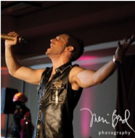 Image of a person in a black vest singing into a microphone with his hands in the air.