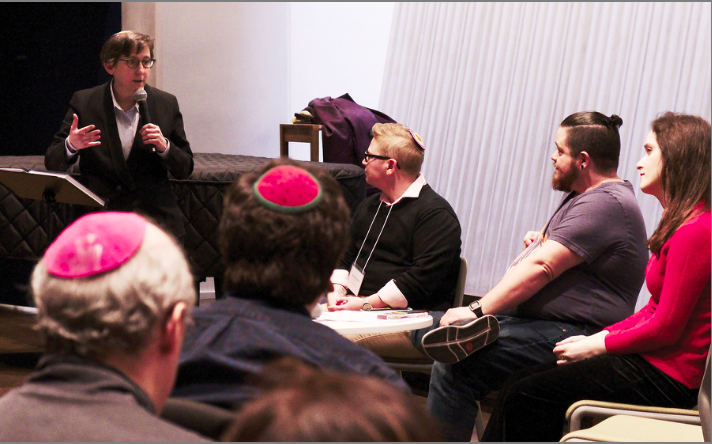 Image of a person speaking into a microphone in front of several others seated in chairs listening attentively.