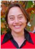 Image of Idit Klein smiling. She is wearing a red shirt with a black collar and is standing in front of leaves.