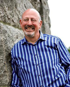 Image of Martin Hannenbaum standing in front of a large rock. He is wearing a blue and white button-down shirt and smiling.