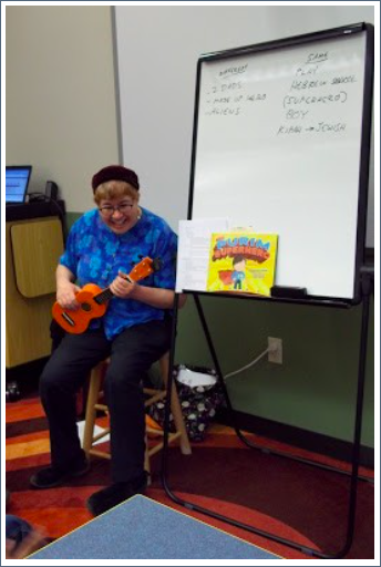Image of Elisabeth Kushner sitting on a stool playing a ukulele. A copy of "The Purim Superhero" sits next to her on a whiteboard. 