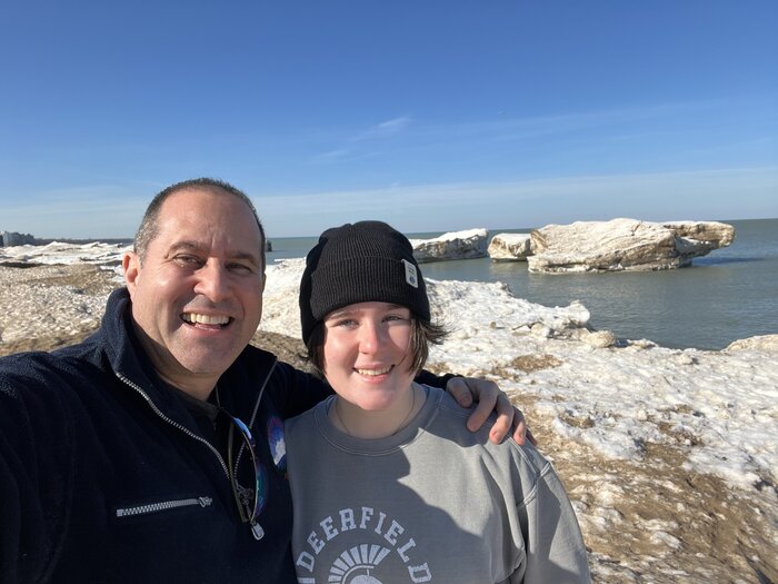 A middle-aged man with short hair with his arm around the shoulder of a teenage boy in a black hat, on a snowy beach