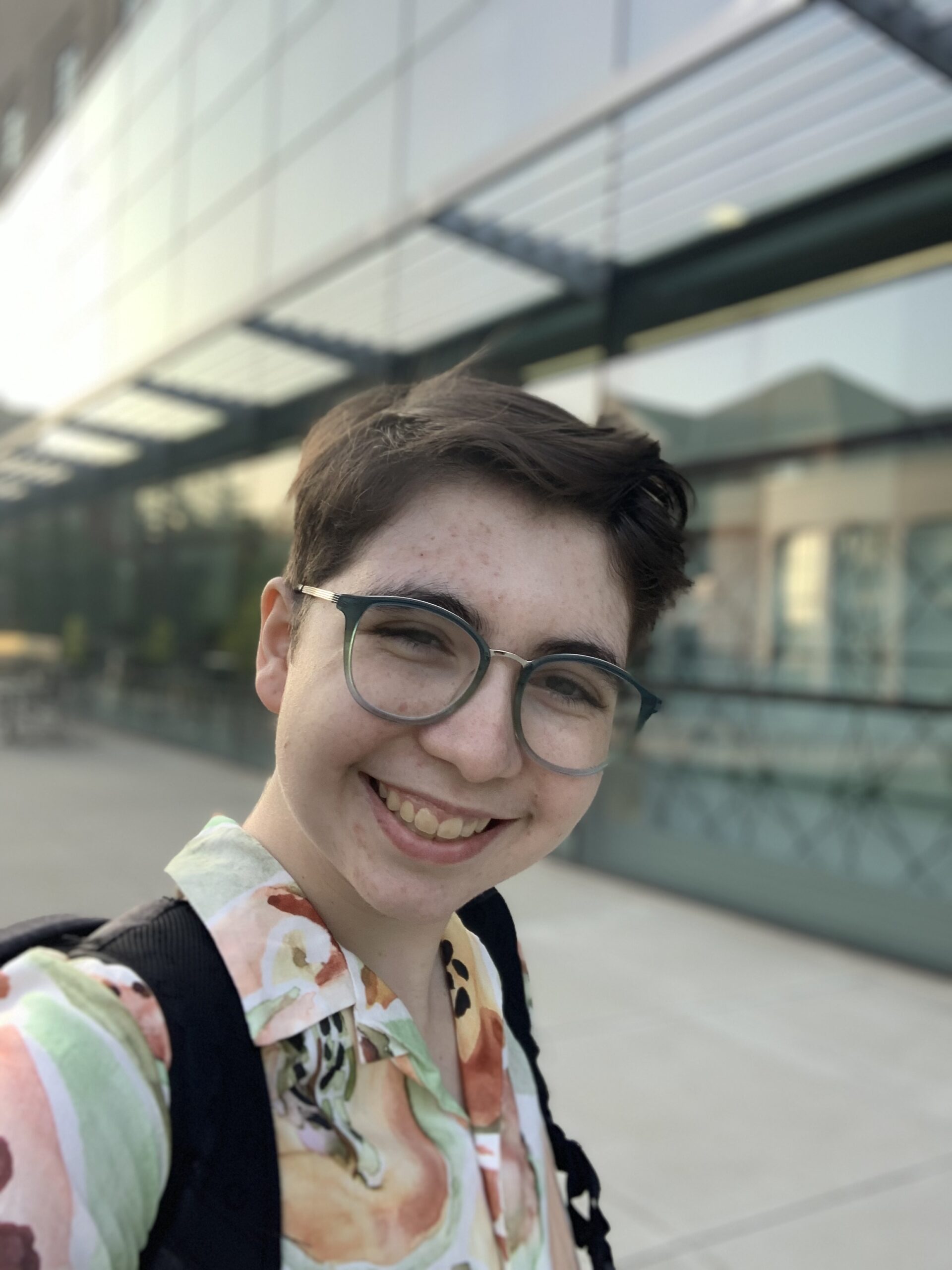 Image of a person with short brown hair and green glasses smiling looking into the camera. They are wearing a colorful button up shirt and a black backpack. They are standing in front of a glass building.