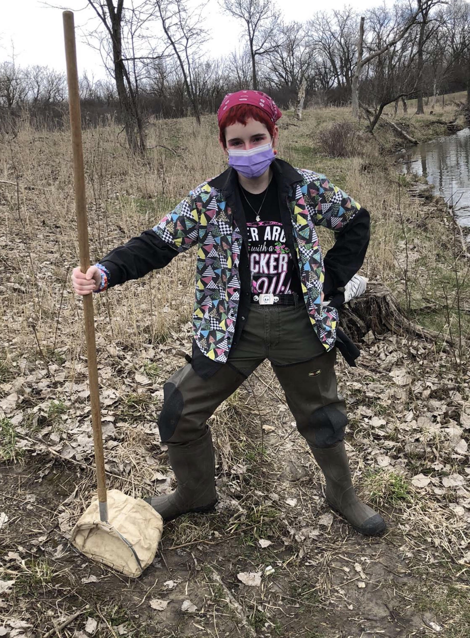 Image of a person in the woods holding a tool and standing with his hand on his hip. He has short red hair and is wearing a red bandanna over it. He is also wearing a purple surgical mask, a colorful button up shirt over a long sleeve shirt, and green pants with mud boots.