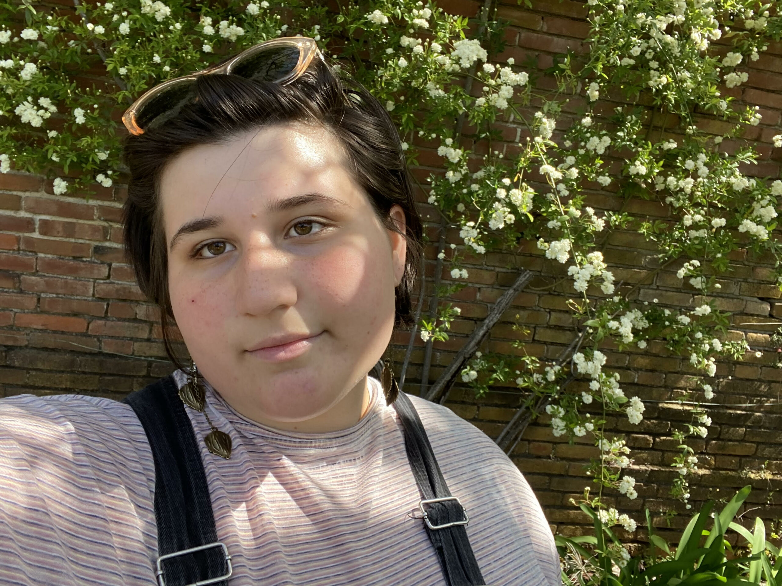 Image of a person smiling. They have straight brown hair and glasses on their head. They are standing on front of a brick wall covered in white flowers.