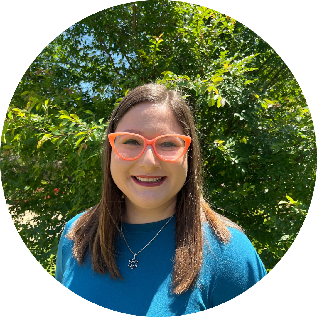 A woman in her 20s with medium light brown hair wearing a cerulean shirt and orange cat eye glasses standing in front of trees